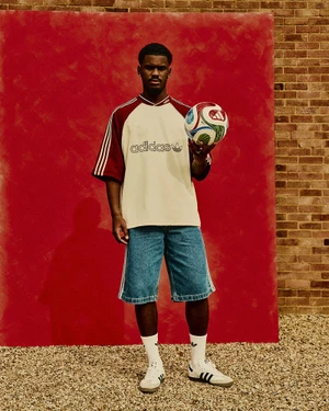 Black man holding a soccer ball while wearing a branded jersey, shorts, and sneakers against a red backdrop.