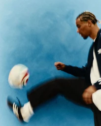 Man with braided hair kicking a soccer ball mid-air with motion blur against a bright blue studio backdrop.