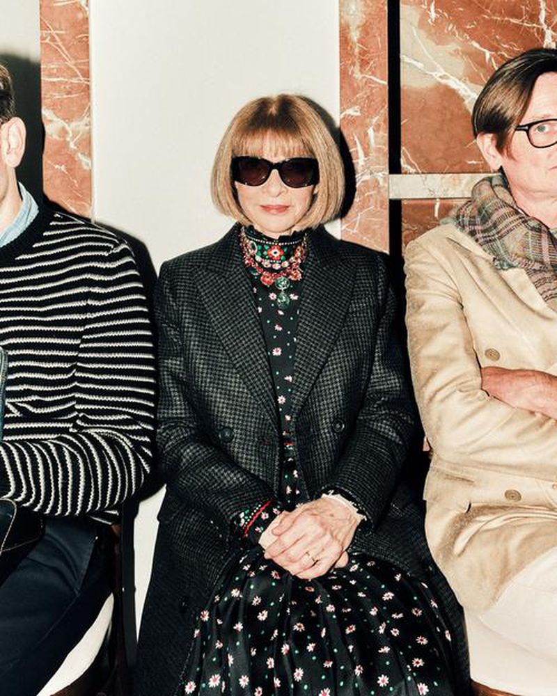 Three people—two men and a woman—are sitting indoors against a marble background. The woman is centered, wearing sunglasses and a patterned dress.