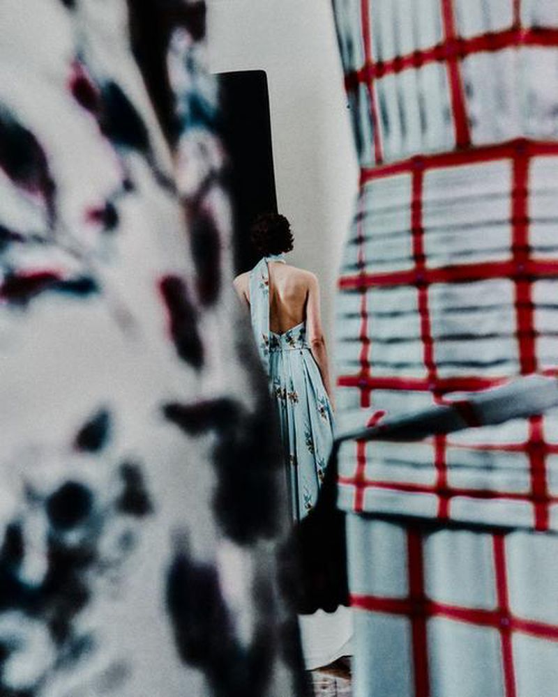 Model seen from behind wearing a light blue halter dress framed between patterned garments during Emilia Wickstead backstage fashion
