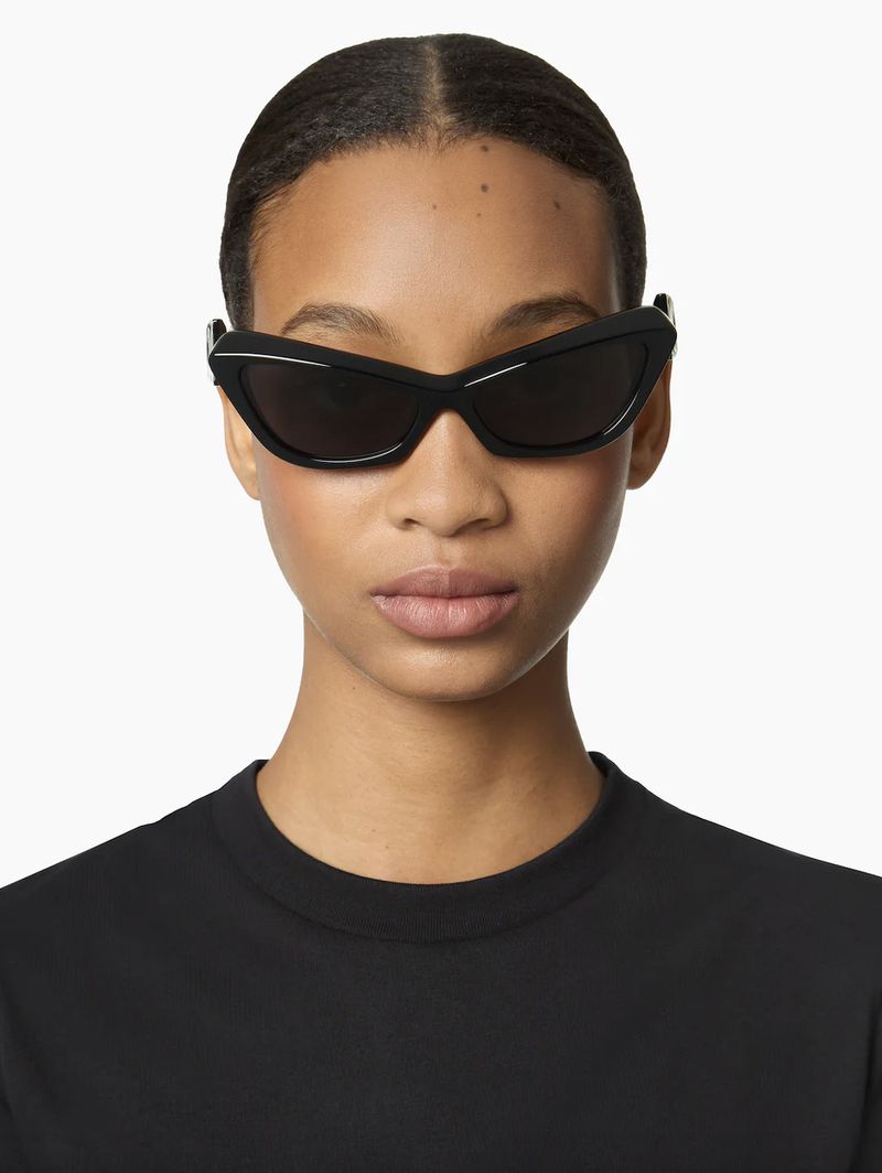 A woman with dark hair wearing black sunglasses and a black shirt poses in front of a white backdrop. The sunglasses are a sharp, angled style.