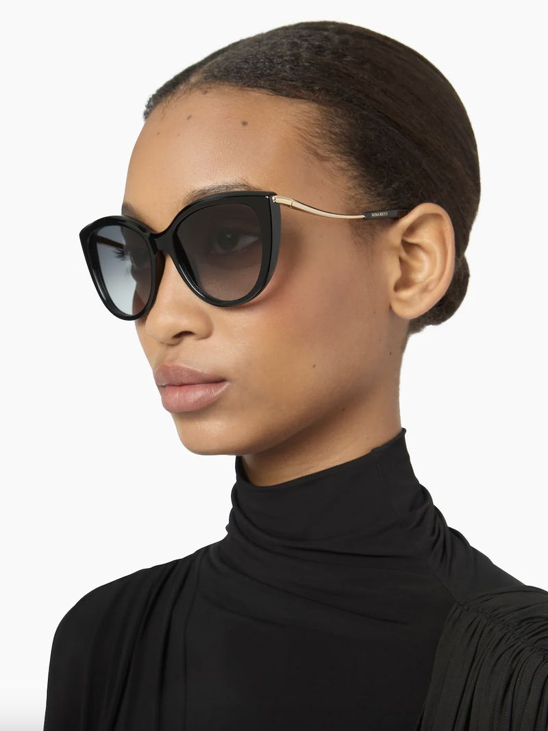 Woman with dark hair wearing black sunglasses and a black turtleneck in a studio shot with a white background.