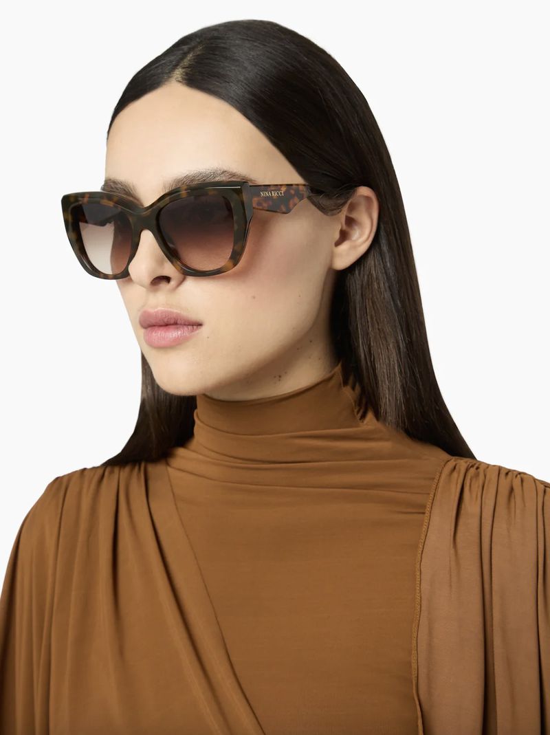 A woman with dark hair wears tortoise-shell sunglasses and a brown turtleneck top, set against a clean white backdrop in a studio shot.