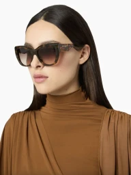 Woman wearing tortoiseshell sunglasses and a brown high neck top against a seamless white background.