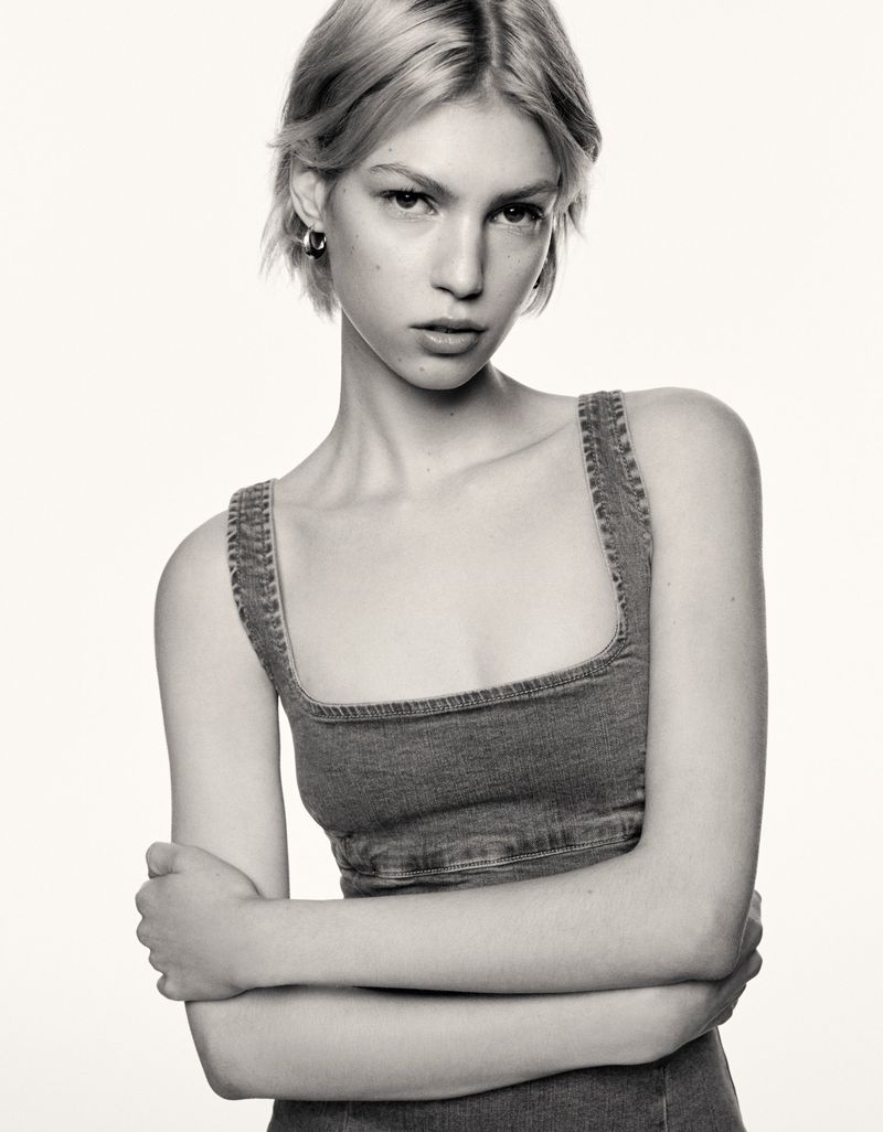Monochrome portrait of a young adult female model in a denim top with crossed arms, posing against a plain backdrop in a photo studio.