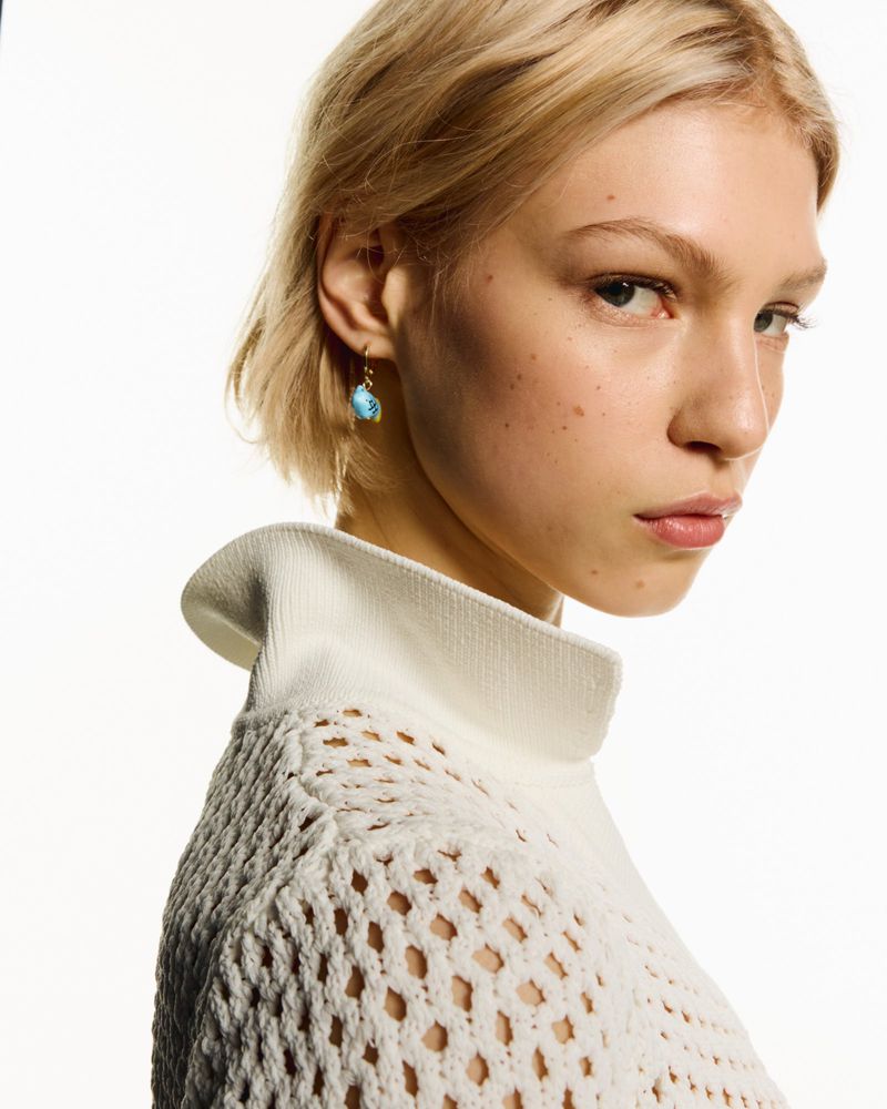 Close-up of a young fair-skinned woman with blonde hair wearing a white knit sweater and turquoise earrings, posing in a studio.