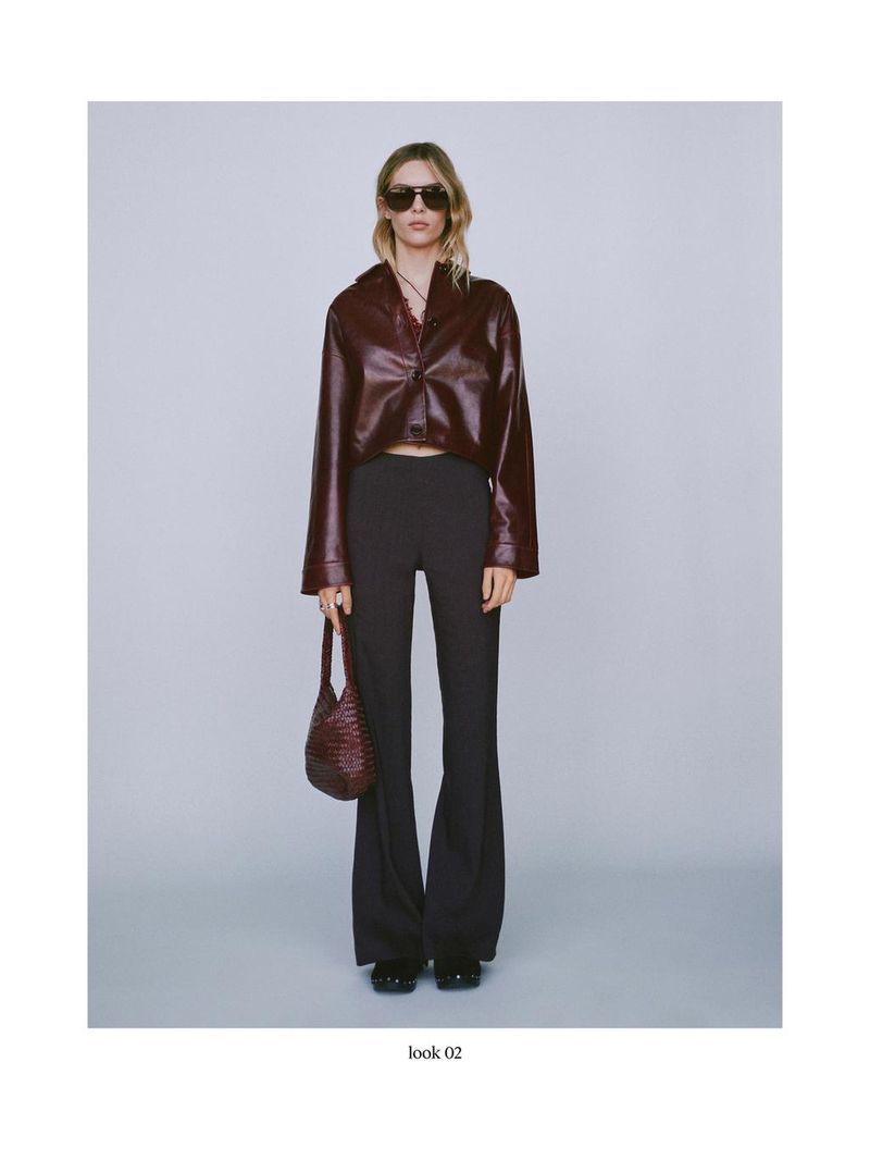 Full-length shot of a woman in a brown faux leather shirt, black pants, and sunglasses holding a woven handbag against a grey background.