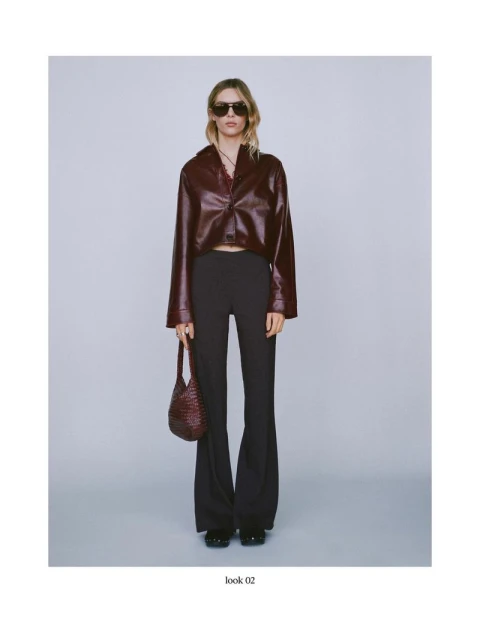 Full-length shot of a woman in a brown faux leather shirt, black pants, and sunglasses holding a woven handbag against a grey background.