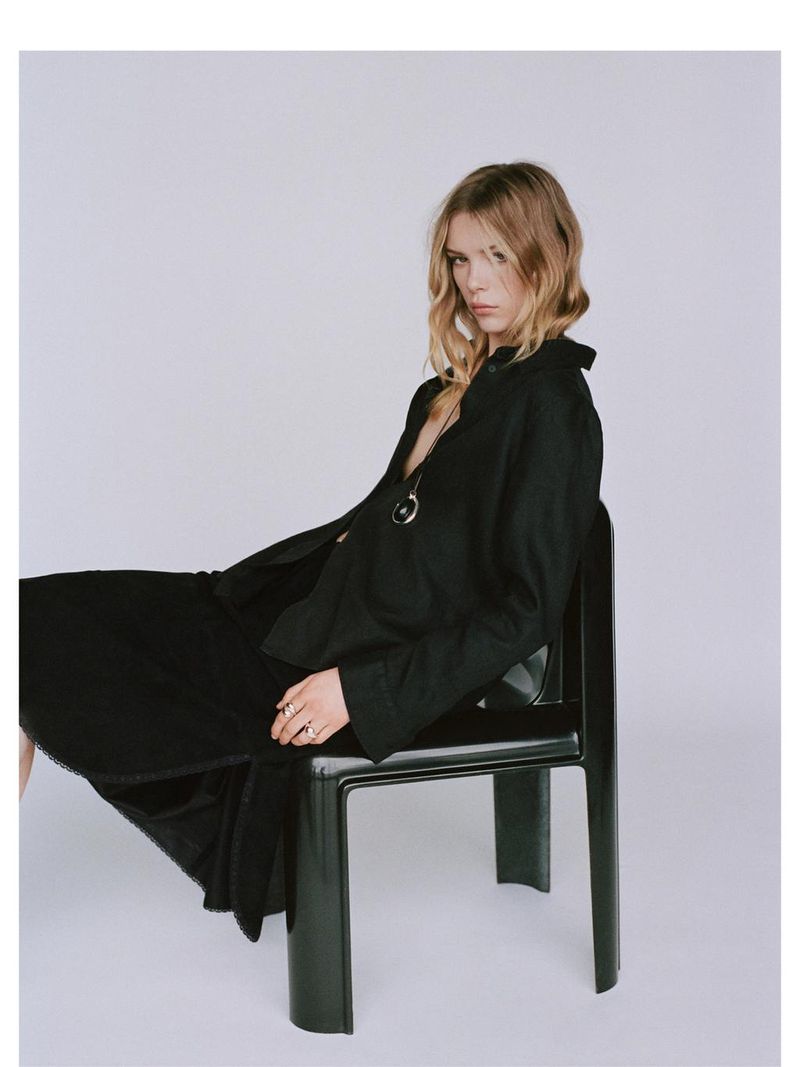 Blonde woman in black dress sits on a chair in a photo studio. She is wearing a silver necklace and has a neutral expression.