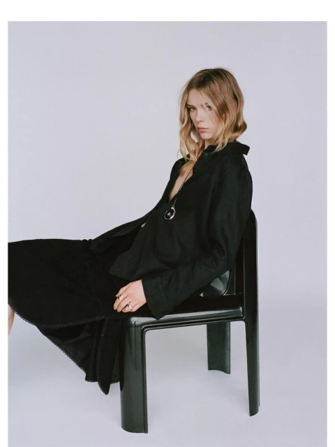Blonde woman in black dress sits on a chair in a photo studio. She is wearing a silver necklace and has a neutral expression.