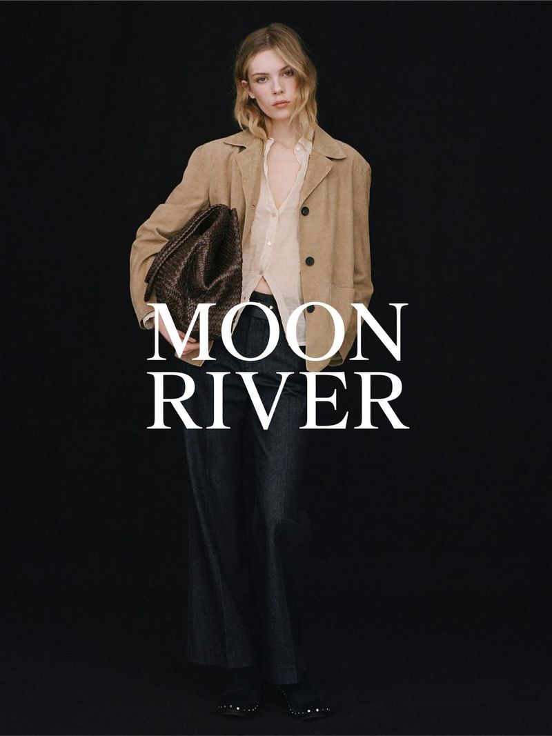Fashionable woman in tan jacket, dark pants, holding a dark brown woven tote bag on black backdrop, advertising 'Moon River' brand.