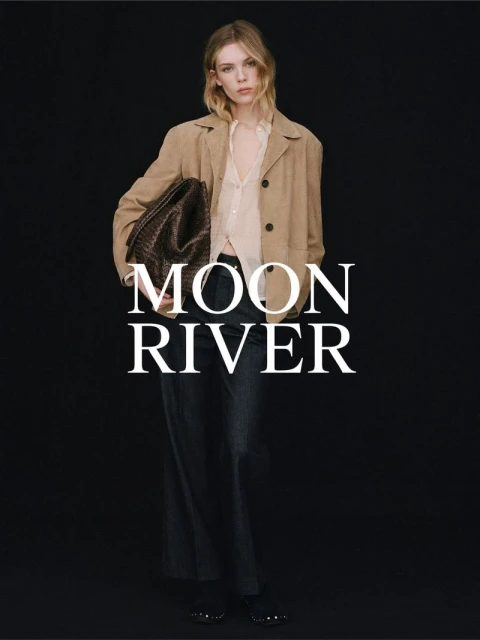 Fashionable woman in tan jacket, dark pants, holding a dark brown woven tote bag on black backdrop, advertising 'Moon River' brand.
