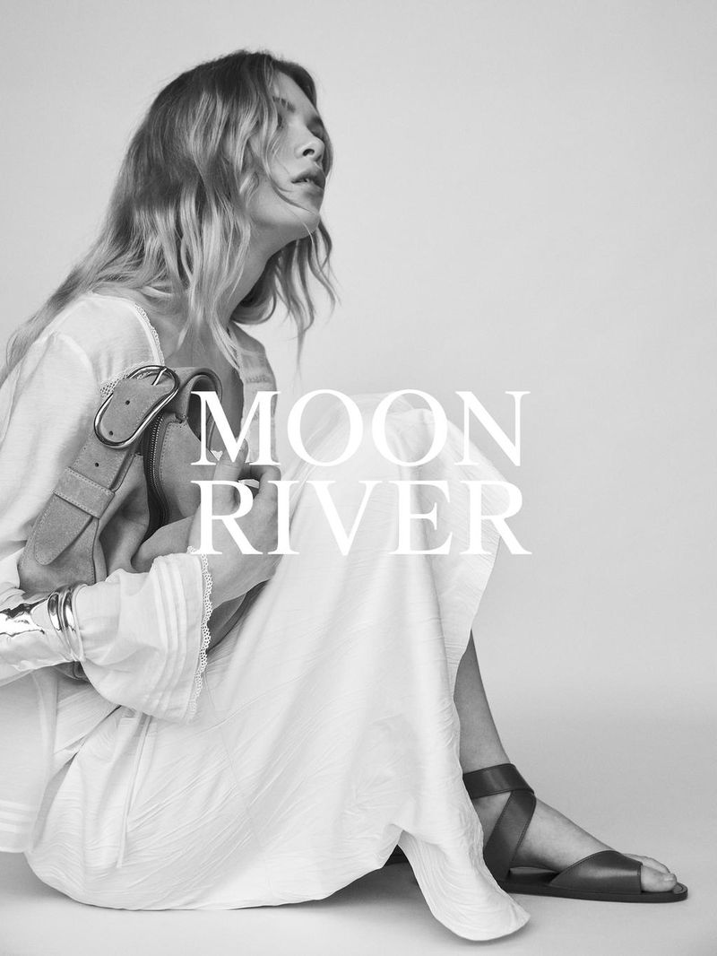 A woman in a flowing white dress sits in a photo studio holding a bag, photographed in black and white, with brand text overlayed.