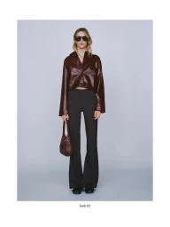 Model wearing a glossy burgundy shirt and dark trousers holding a matching handbag against a grey seamless studio background.