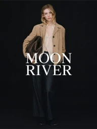 Model in camel jacket and dark trousers holding a large woven clutch against a plain black background for Massimo Dutti