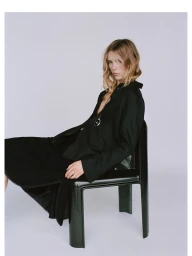 Woman in a black shirt and long skirt seated on a dark chair against a pale grey studio background