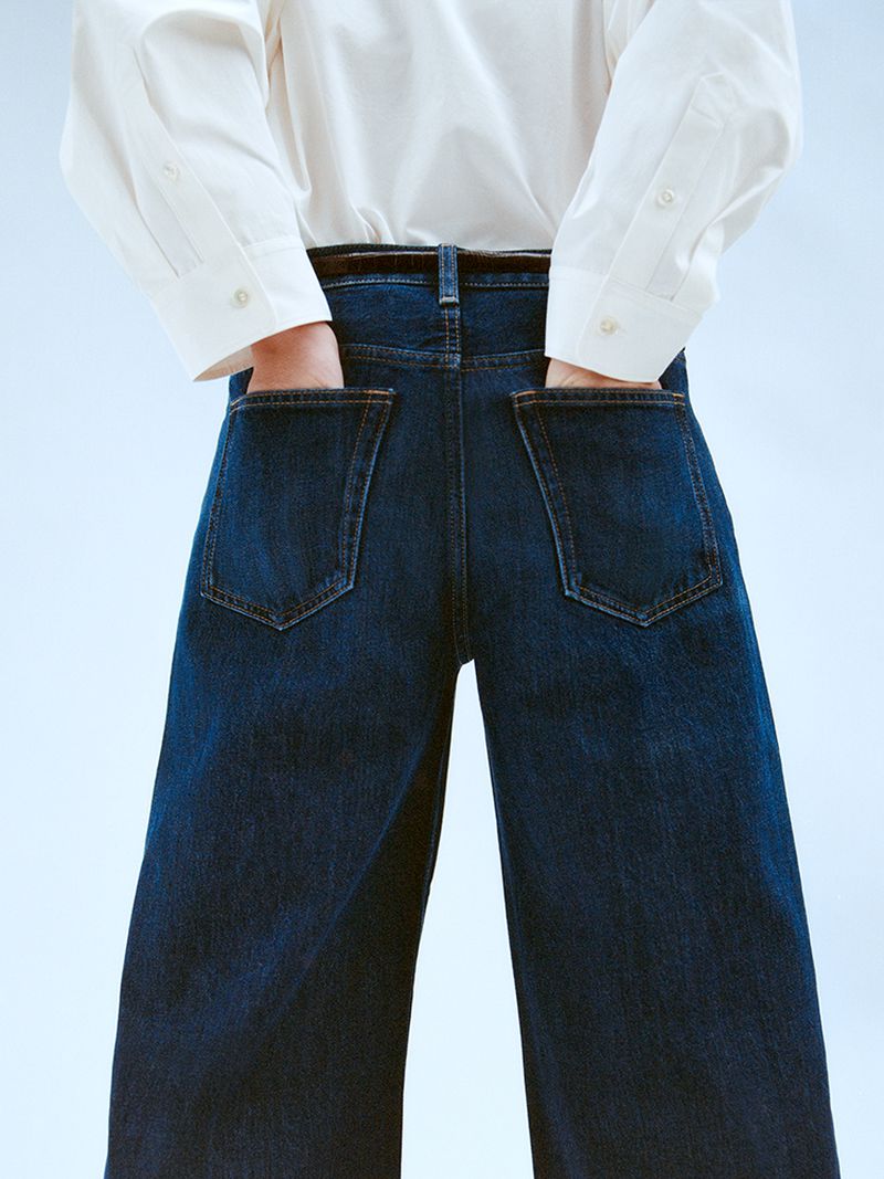 Person wearing dark wash wide-leg jeans and white button-down shirt with hands in pockets against a soft gradient background.