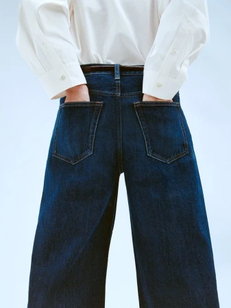 Person wearing dark wash wide-leg jeans and white button-down shirt with hands in pockets against a soft gradient background.