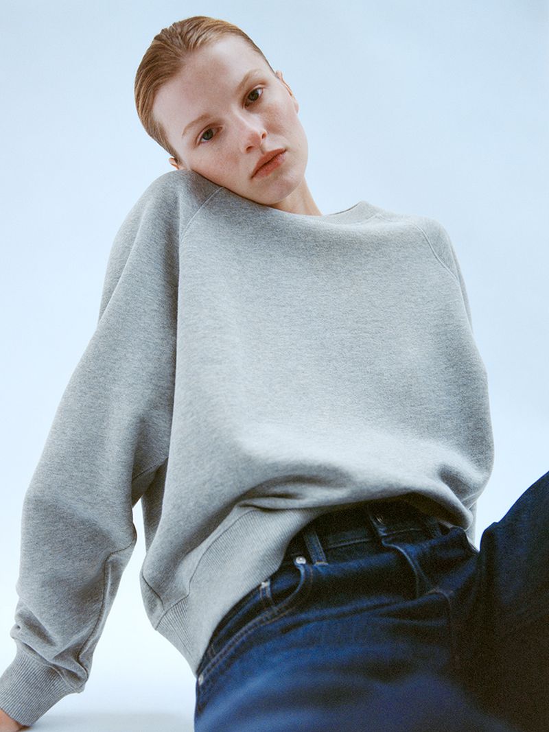A model poses in a studio wearing a grey sweatshirt and dark jeans, captured in soft lighting with a light blue backdrop.