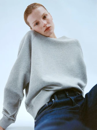 A model poses in a studio wearing a grey sweatshirt and dark jeans, captured in soft lighting with a light blue backdrop.