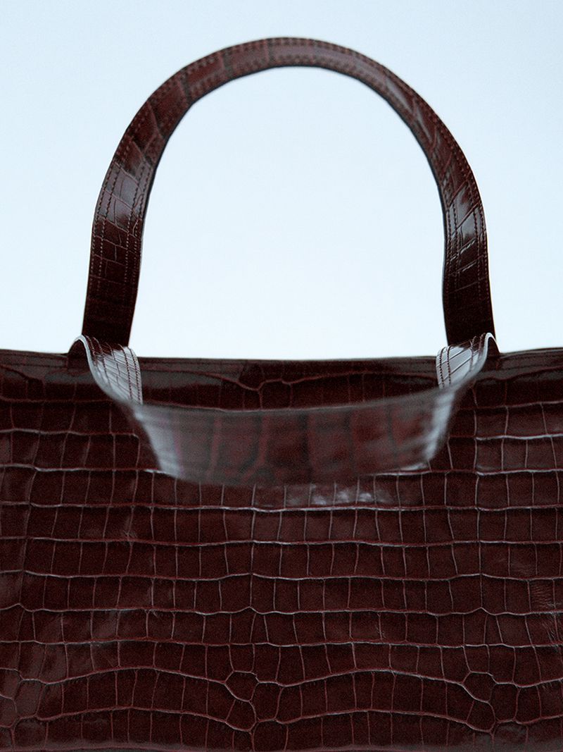 Cropped shot of a brown, textured, leather handbag against a pale blue background in a studio setting with bright, even lighting.
