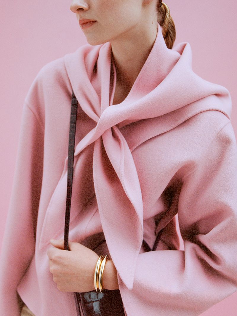 Woman in pink coat with stylish tie detail, accessorized with gold bracelets and a dark brown handbag against a pink backdrop.