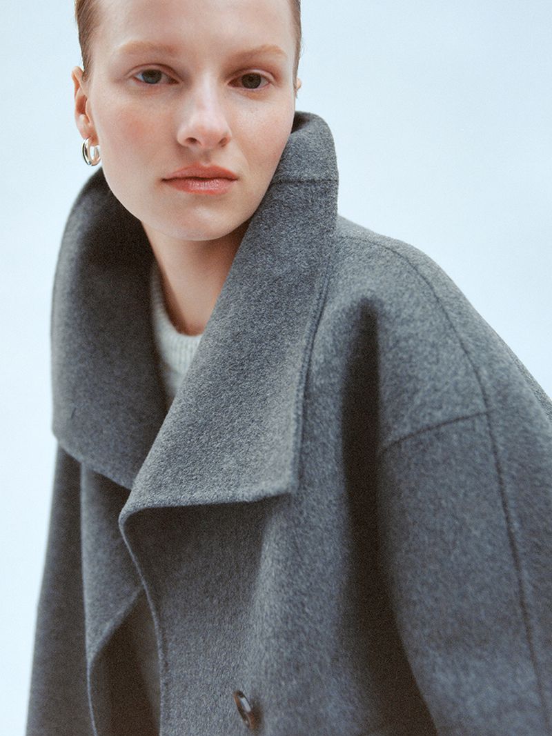 Close-up of a stylish adult female in a grey wool coat. The portrait showcases a high-fashion look with natural light, indoors.