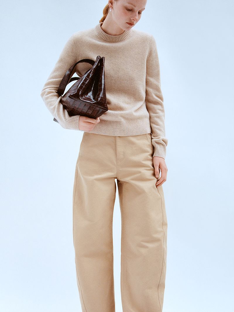 A fair-skinned woman in a beige sweater and wide-leg tan pants holds a dark brown crocodile-skin tote bag in a studio setting.