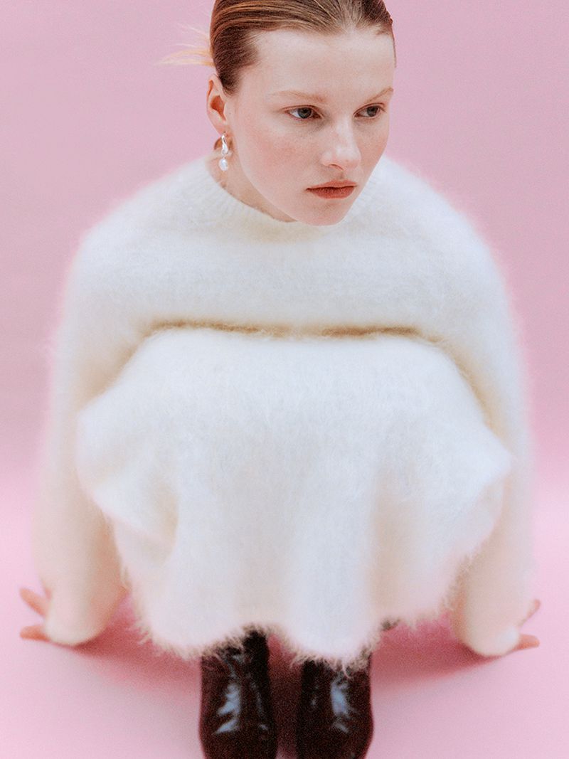 Fashion shot featuring a model in a fluffy white sweater and dark patent leather shoes, posing against a pastel pink backdrop.