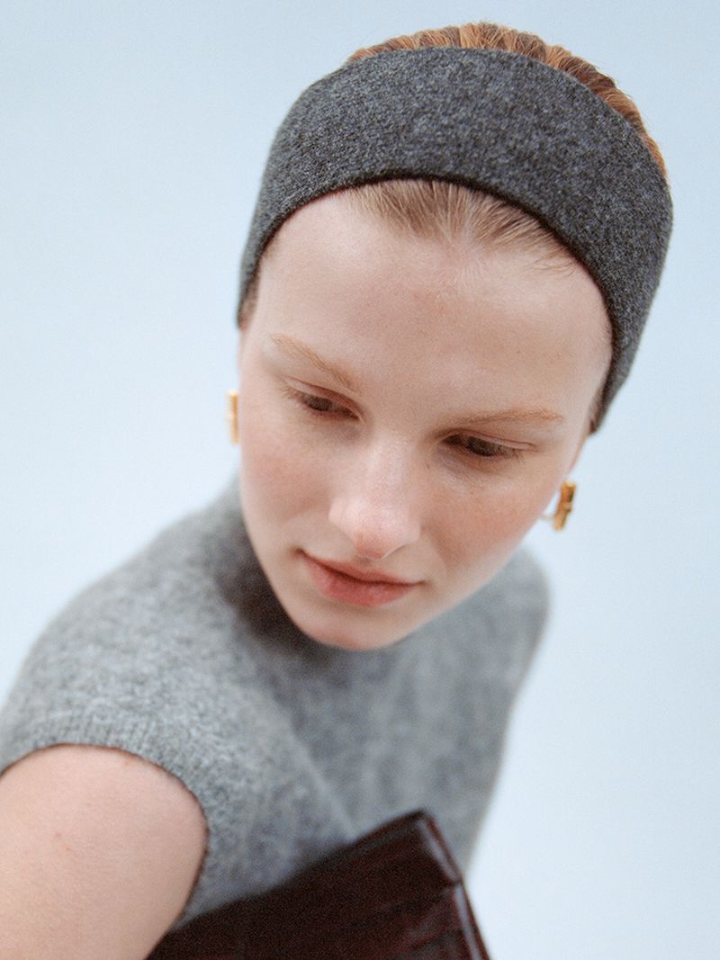 Close-up portrait of a fair-skinned woman wearing a grey headband and grey sleeveless top, accessorized with gold square earrings, indoor lighting.