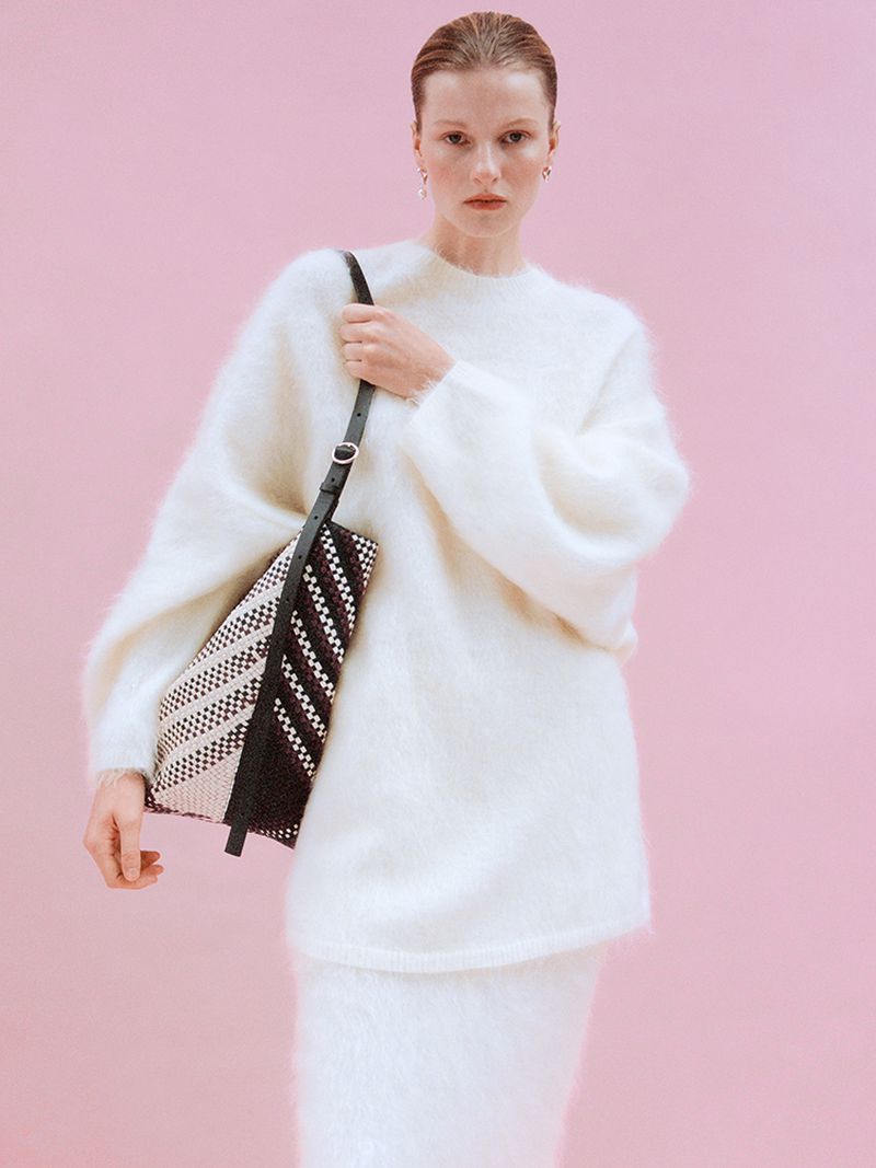 Model in a fluffy white sweater holding a patterned woven handbag against a pale pink seamless backdrop for Arket.