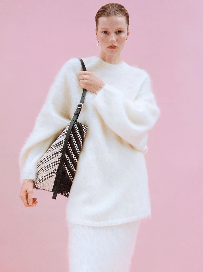 Model in a fluffy white sweater holding a patterned woven handbag against a pale pink seamless backdrop for Arket.