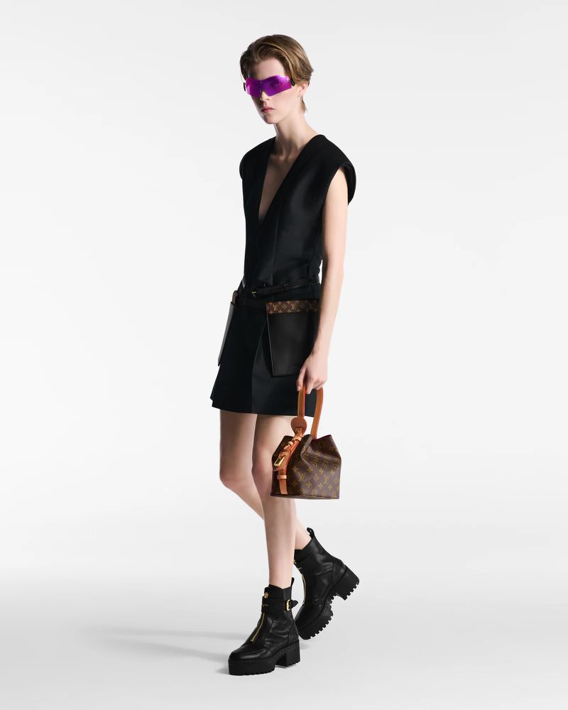 Fashion model posing in a black dress and boots holding a monogram handbag in a studio. Purple visor-like sunglasses. White background.