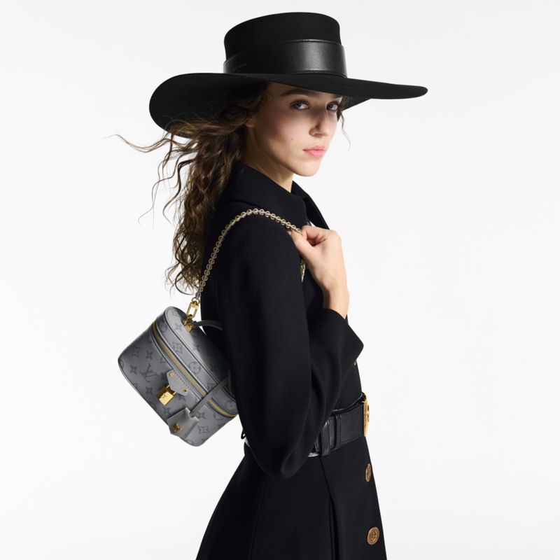Elegant woman in black hat and coat with a designer purse, posing in a studio with a clean white backdrop, highlighting high fashion.