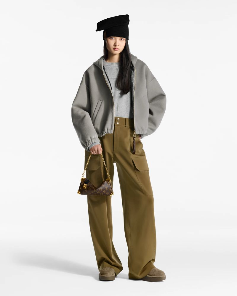 A slender Asian fashion model in a grey jacket and olive green pants holds a patterned handbag, posing against a clean white backdrop.