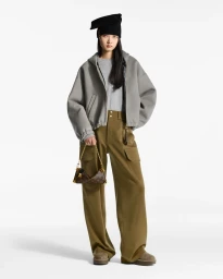 Model wearing a grey zip jacket, olive trousers, and a black hat holding a monogram handbag against a white background