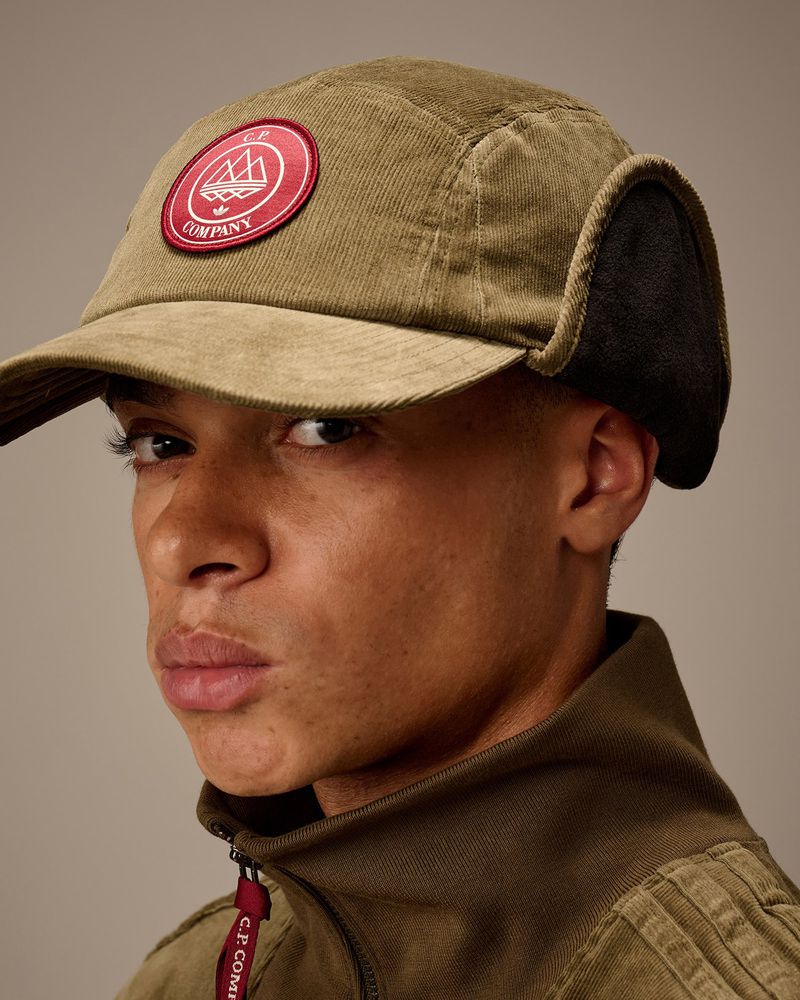 Close-up of a brown-skinned man wearing an olive green corduroy trapper hat with a logo, and a matching jacket, against a neutral background.