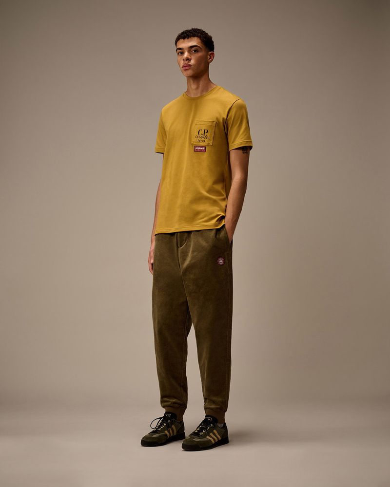 Adult male standing in a photo studio, wearing a mustard-colored shirt, brown sweatpants and Adidas sneakers in a full shot.