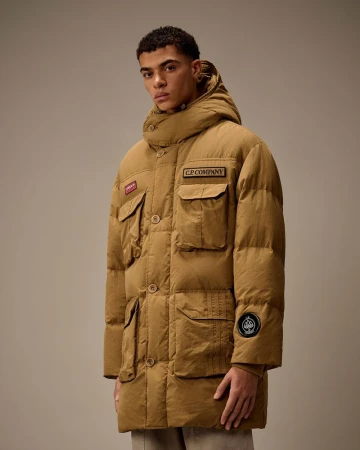 A brown-skinned adult male poses indoors wearing a tan puffer coat with pockets and buttons, complemented by neutral pants and studio lighting.