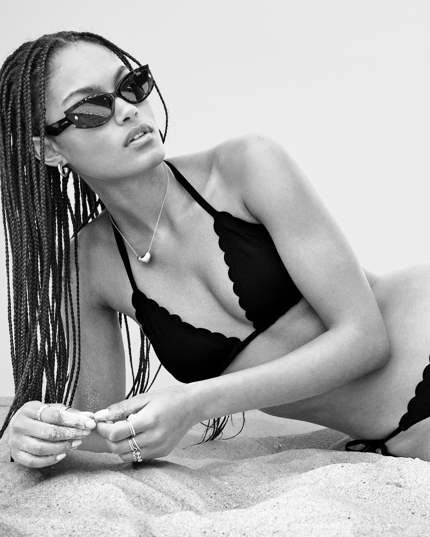 A woman with braided hair is lying on a sandy beach wearing a black bikini and sunglasses in a black and white fashion shot.
