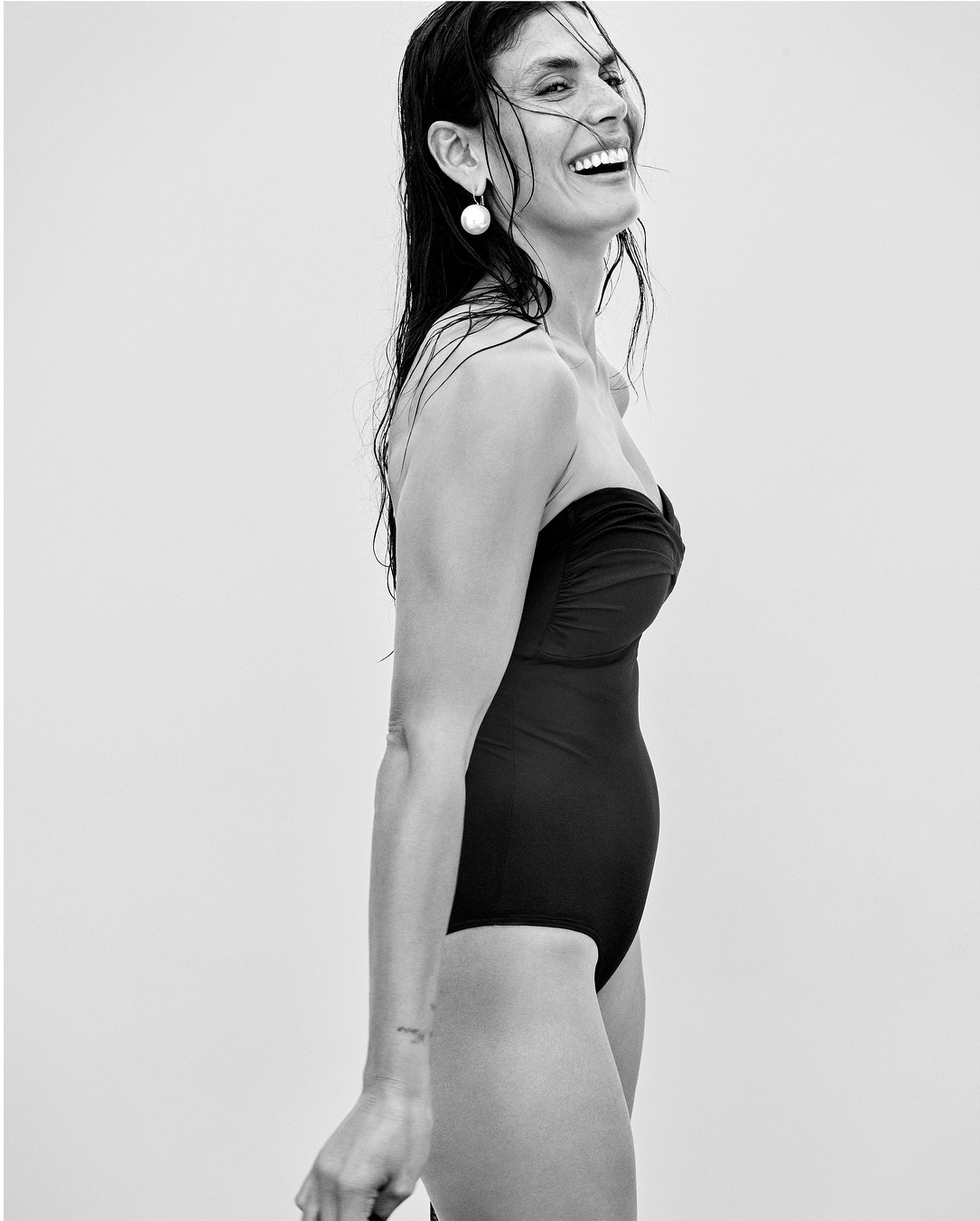 Black and white portrait of a smiling woman with wet dark hair in a black one-piece swimsuit against a neutral background.