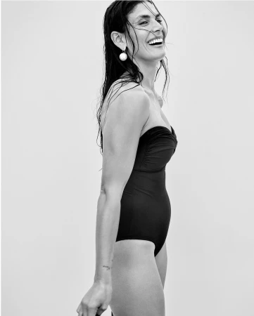 Black and white portrait of a smiling woman with wet dark hair in a black one-piece swimsuit against a neutral background.