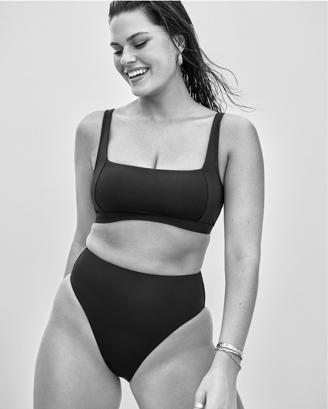A smiling adult woman in a black two-piece swimsuit with natural hair poses against a light backdrop in a studio setting.