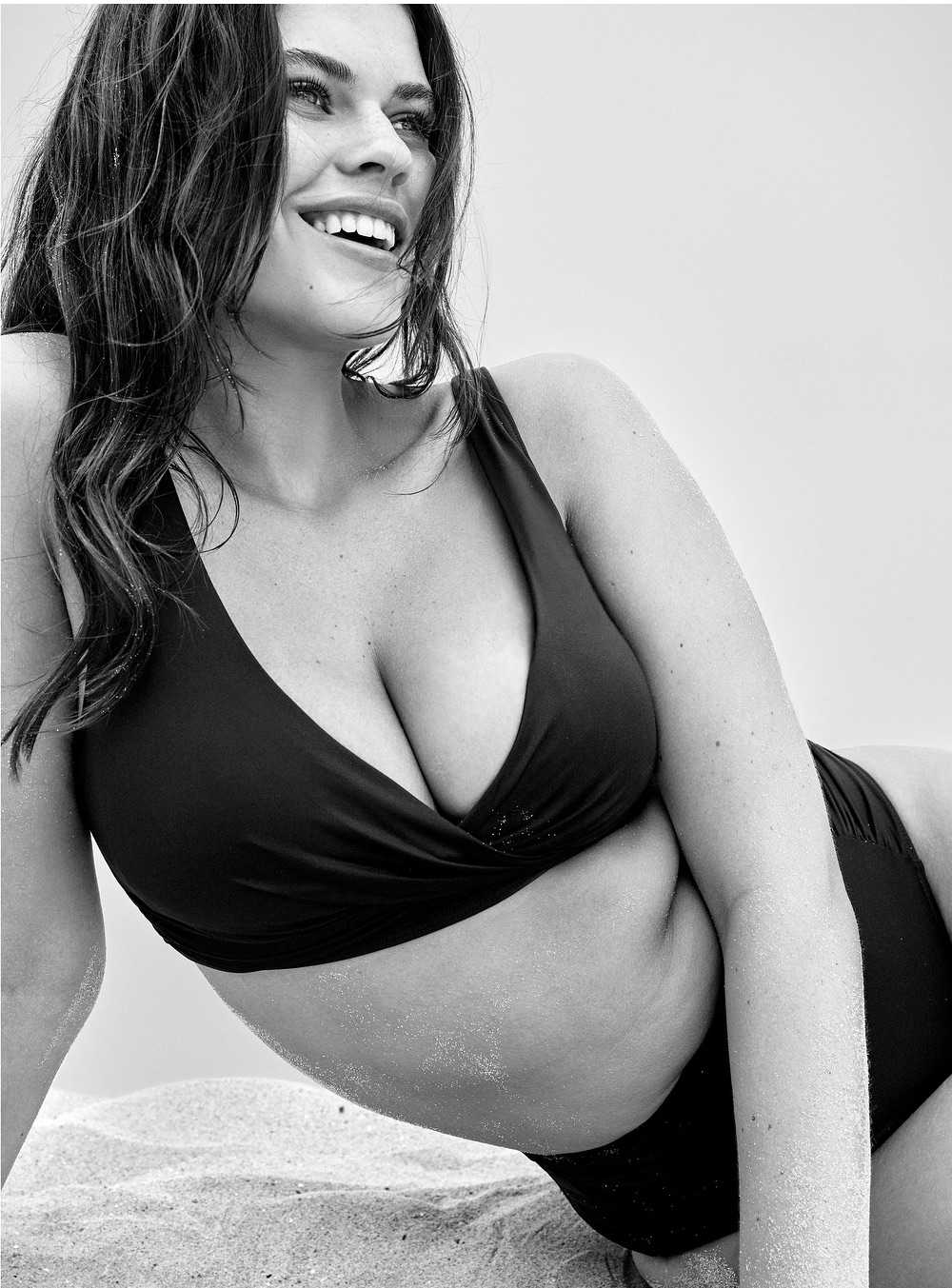 Black and white portrait of a smiling woman in a bikini lying on a sandy surface, close-up shot, evoking a natural, confident mood.