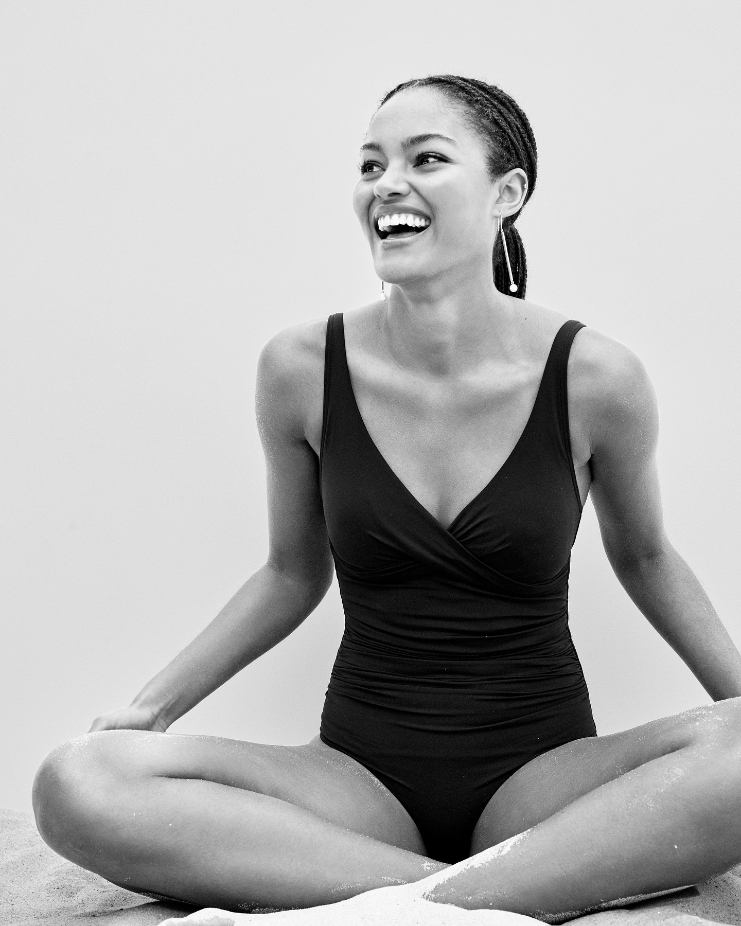 A joyful African American woman in a black swimsuit sits cross-legged on a sandy beach, radiating happiness in a black and white portrait.
