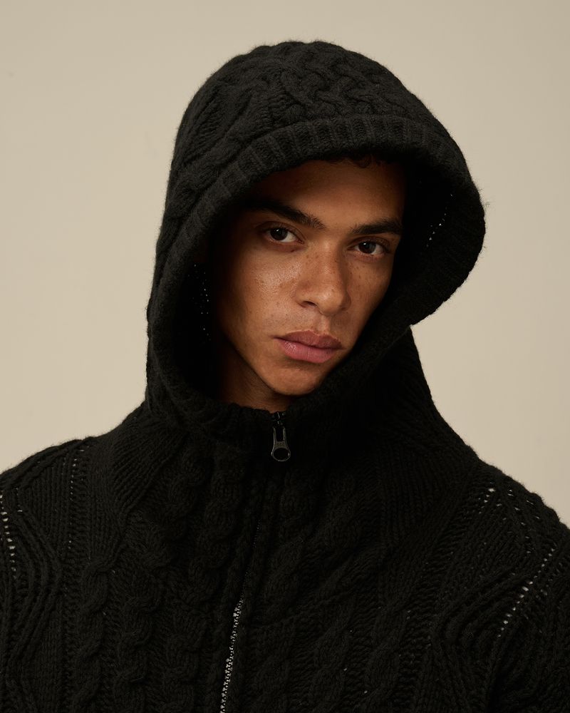 Adult male wearing a hooded, cable-knit wool jacket indoors against a plain background. Close-up portrait with neutral expression.