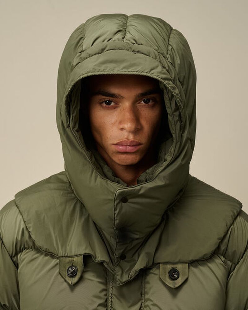 Close-up portrait of a man wearing a puffy olive green hooded jacket against a light beige backdrop, focusing on fashion and portraiture.