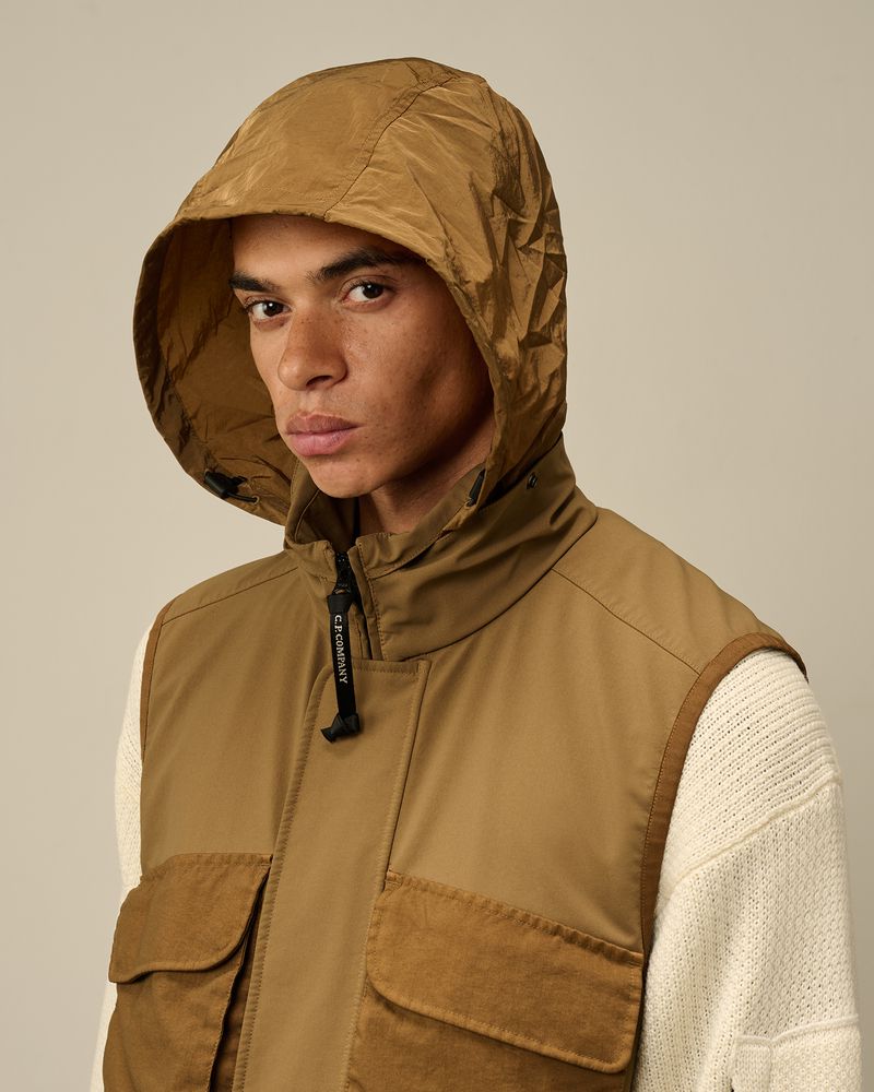 Close-up of a handsome adult male model wearing a hooded tan vest over a cream-colored sweater in a brightly lit photo studio.