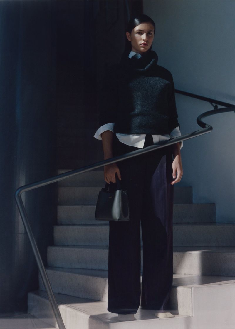 Stylish woman in dark sweater and wide-leg pants stands on stairs with a dark green handbag, bathed in natural light.