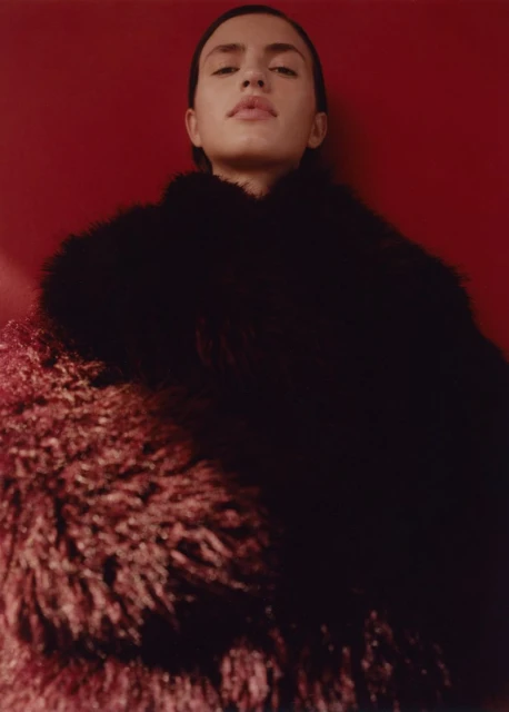 Portrait of a fair-skinned woman with long dark hair, wearing a fuzzy coat, posing against a red backdrop. Fashion photography.